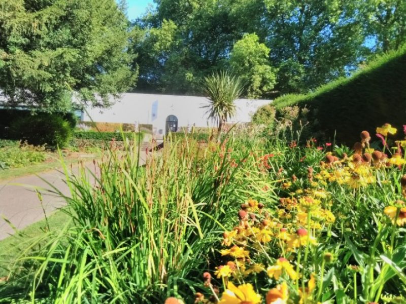 LUX building. A long white building seen in the distance surrounded by trees, in the foreground are flowers and green plants, its a sunny day