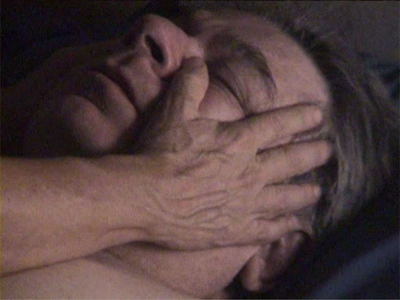 a white man lying on a hospital bed with his eyes closed. A closeup of a pair of hands caressing the man's face.