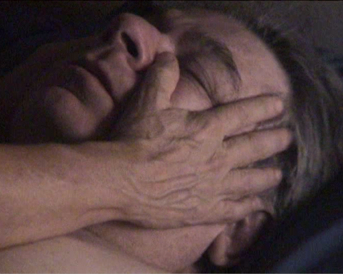 A close up of a head of a white man lying with his eyes closed. A pair of hands caressing man’s face.