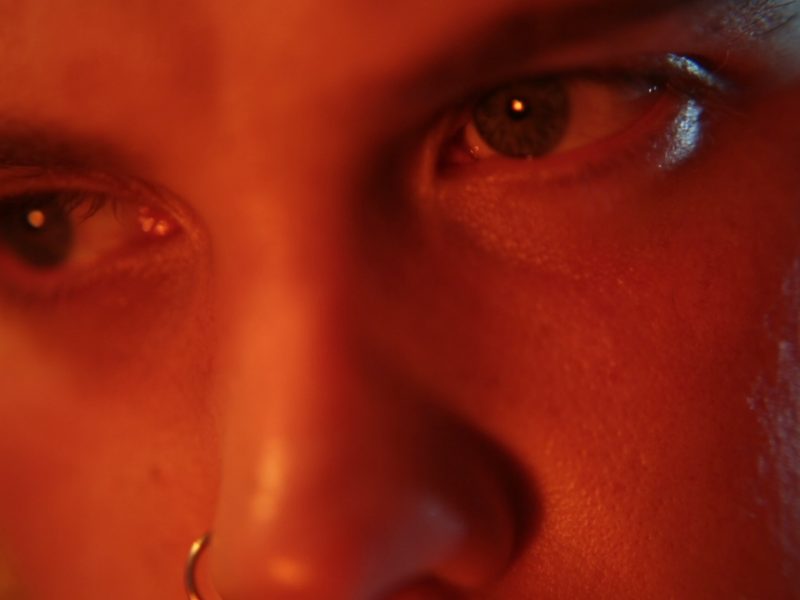 A closeup of a white person’s eyes looking intensely at something outside the frame. A dark orange light hues the face and there is a hint of a blue light coming from behind.