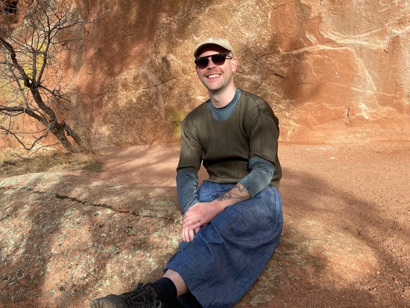 Jordan Lord, a white person with a blonde mustache, smiles at the camera, wearing a hat and sunglasses, with their hands folded over their legs, as they sit on a red rock on a sunny day.