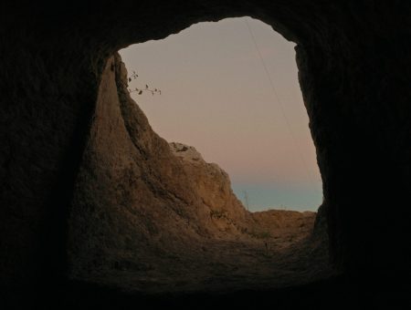 Image from inside a cave looking up and out to the walls of the cave and the sky that is a gradient of grey, to pink, to blue. The grey and pink being clouds, and the blue being the sky.
