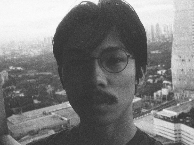 Black and white photograph of filmmaker Miko Revereza standing on what appears to be a balcony overlooking a cityscape. He is wearing round glasses and has a moustache, with his face partially in shadow. Tall buildings and a skyline can be seen in the background.