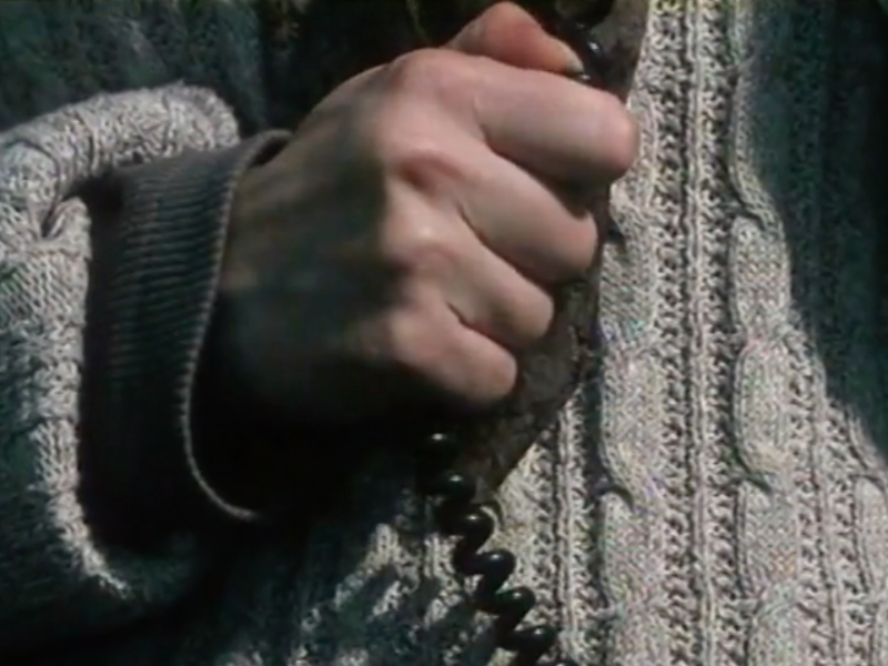 We see a close-up of a hand tightly gripping a telephone cable, held close to the body that is wearing a gray jumper. The hand appears tense, with visible strain on the fingers and knuckles.