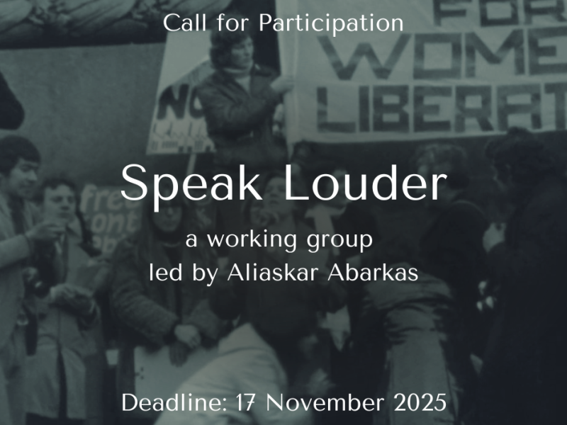 A black and white photograph of a group of protestors wrapped up warmly in winter coats, holding placards. On a raised platform above stands a person holding part of a large tapestry that reads 'Womens Liberation'.