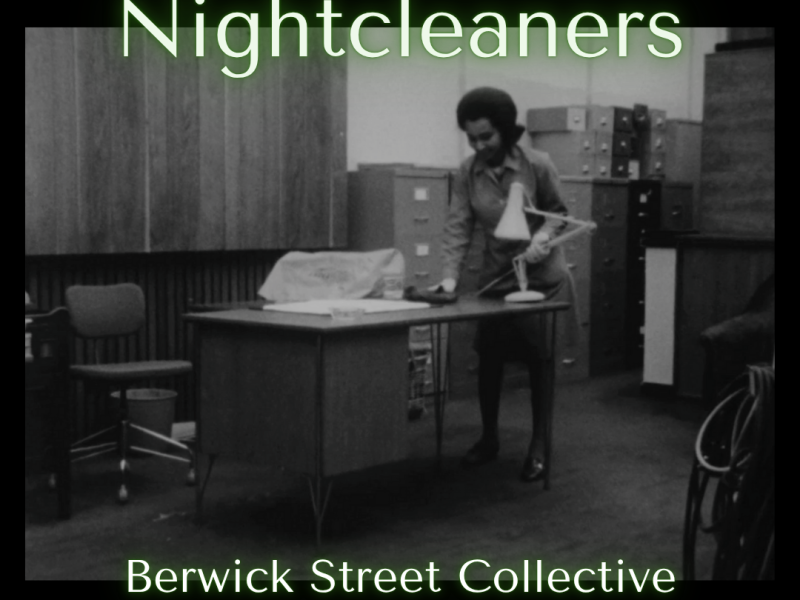 A black and white photograph of an office furnished with rows of paperwork cabinets. In the foreground stands a large wooden desk, behind which a person is wiping its surface with a cloth.