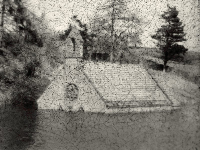 A house submerged in a river, with only the roof visible, set against a backdrop of trees. The surface of the image is heavily crackled.