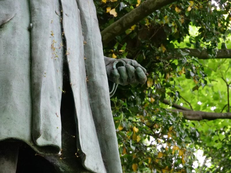 a detail of statue of Sir Sidney Waterlow, Waterlow Park, Highgate, in which can be viewed a frock coat and hand holding a pocket watch in grey stone with green leaves in the background.