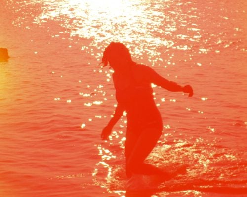 A washed out reddish image of a woman in the water with the sun shining, a film still from Pyramid by Margaret Salmon