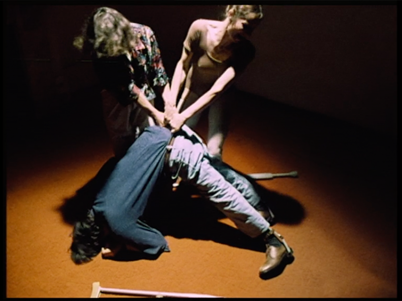 Two women are pulling a man’s waist up while their upper body creates V-shape. The man’s head and feet are on the floor creating a reversed V-shape. a pair of crutches fell on the orange floor.