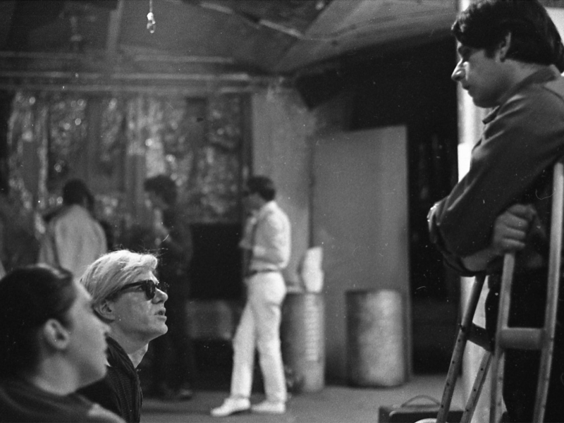 a black and white image, on the left hand side Andy Warhol with white hair and dark glasses looks up at Stephen Dwoskin who stands resting on a pair of crutches looking down at him