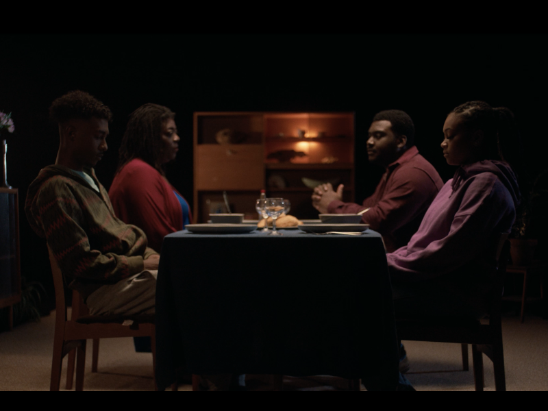 In a sound stage with black walls, a family of four individuals with dark skin is seated around a table. They wear solemn expressions, illuminated by soft, dim lighting. The warm glow of lamp lights highlights the wooden furniture in the background
