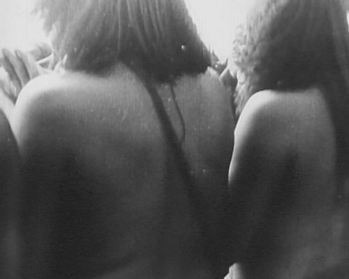 The backs of naked black women standing in a row. A face of a black man in front of them is seen at the left top corner.