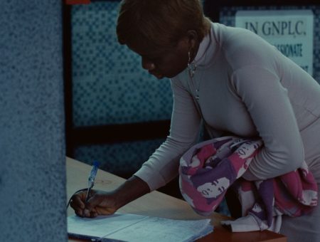 Pitcture of a person in a grey dress hunched over writing something on a piece of paper with one hand and holding a sweater.