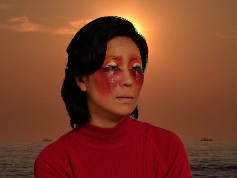 A Korean woman is wearing face paint around her eyes that resembles a sunset against the backdrop of the ocean during sunset