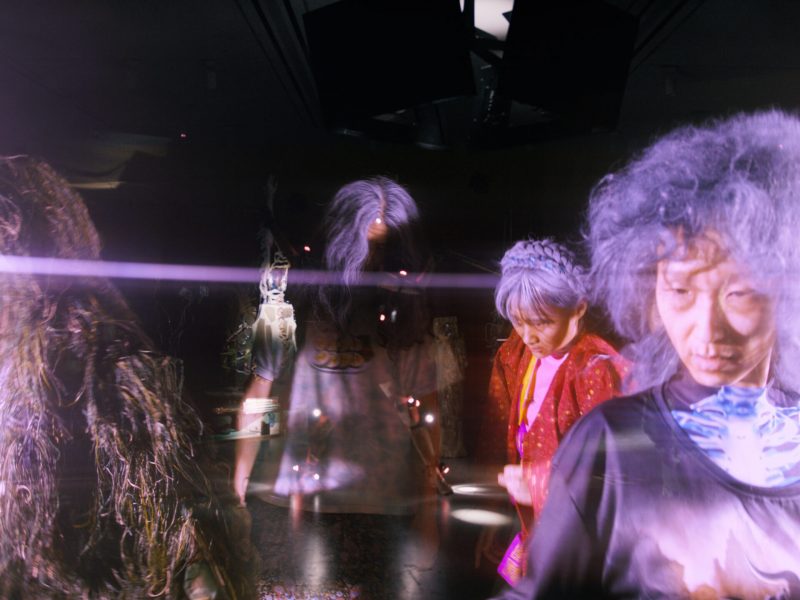 A dark empty gallery is overlaid with a ghostly images of four performers in old age makesup and wigs with blank stares.