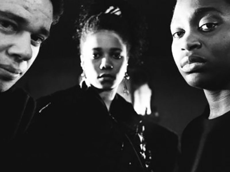 Three people, a white man and two black women, looking directly into the camera under a dramatic lighting creating a film noir look.
