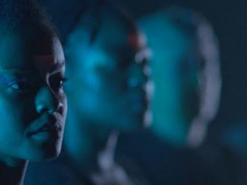 Blue light filled in a dark space is reflected on the face of a black woman in a foreground. Two other people in the background are blurry.