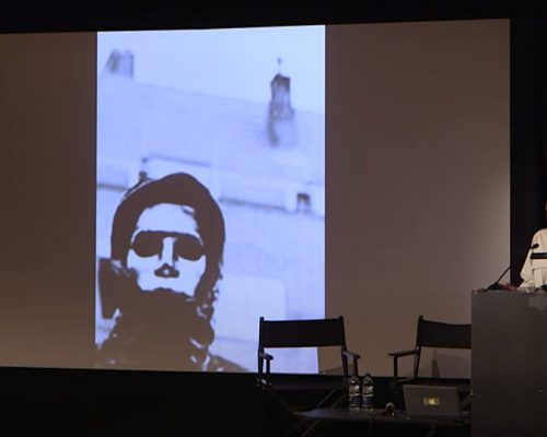 A black person is reading in front of a speaker stand on a stage. A large screen is projecting an image of a person in a full face mask.