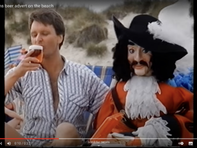 image from McEwans Advert. A man is drinking a glass of beer, next to him is a puppet cavalier, an embodiment of the McEwans logo
