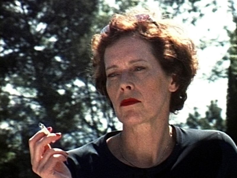 A white woman wearing red lipstick is holding a cigarette and gazing blankly. The edges of her curly hair shine from the bright sunlight coming through trees behind her whiles her face is in shadow.