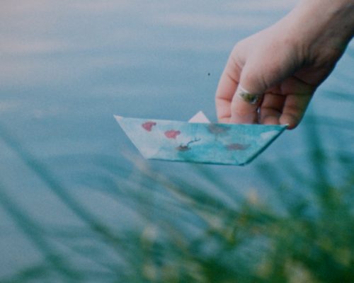 A hand gently holds a small paper boat decorated with blue and red drawings. The boat hovers just above the surface of a calm river, with green grass in the foreground.