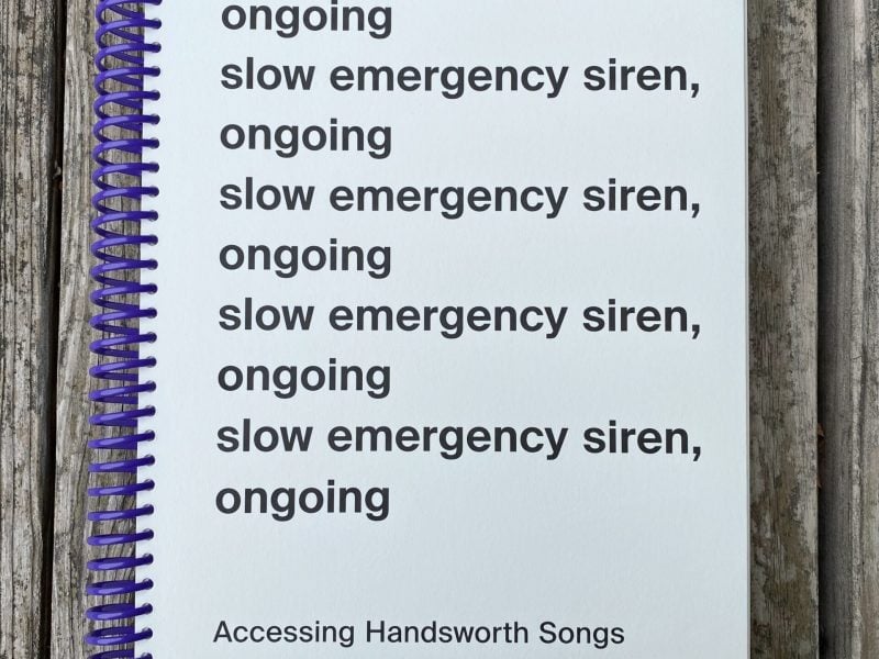 a white book with a large purple spiral binding placed on a wooden surface. On the cover is the title slow emergency siren, ongoing written five times and below the title is subtitles that read accessing handsworth song edited by Sarah Hayden