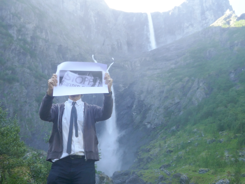 A person holding a piece of paper with a word ‘STOPP’ printed in black and white, obscuring the head of the person. Behind the person is a mountain range with two waterfalls
