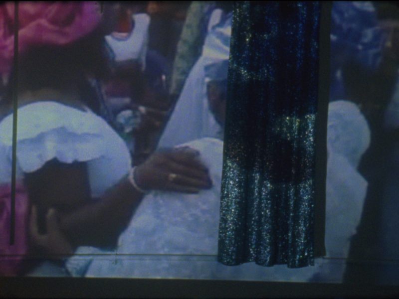 An image of black women in white voluminous dresses with frills and colorful headpieces is projected onto a white wall and a glittery curtain drape.