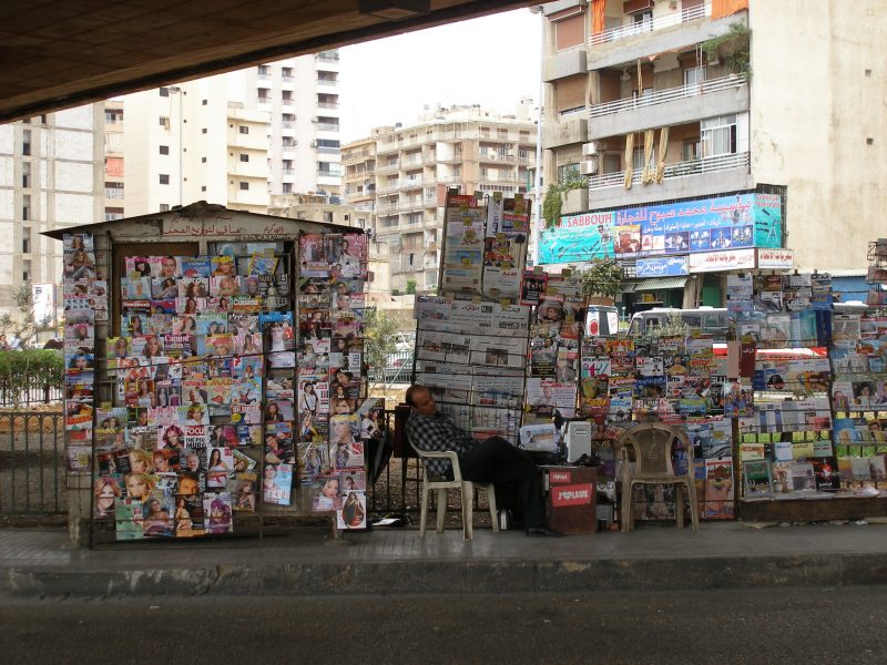 image from the film An Untimely Film for Every One and No One First and Second Part (2018), an image of a newspaper seller under an underpass, there are many colourful magazines being displayed, and a man dozes in a chair in the centre of the image, behind are some buildings