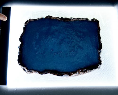 On a glowing led lightbox is a ceramic tray with erratic edges filled with blue liquid. A finger wearing a silver ring touches the power button on the lightbox